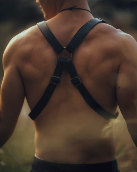 Back view of man wearing Bijou by Juju black leather suspenders with brass hardware in a natural outdoor setting.